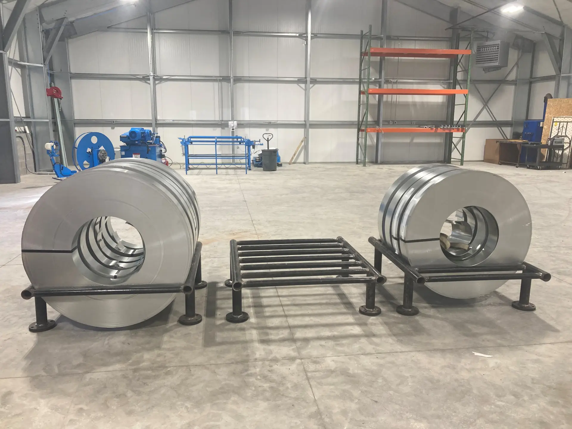 Two stacks of large steel coils sit on black metal stands in a spacious industrial warehouse with machinery and shelving in the background.